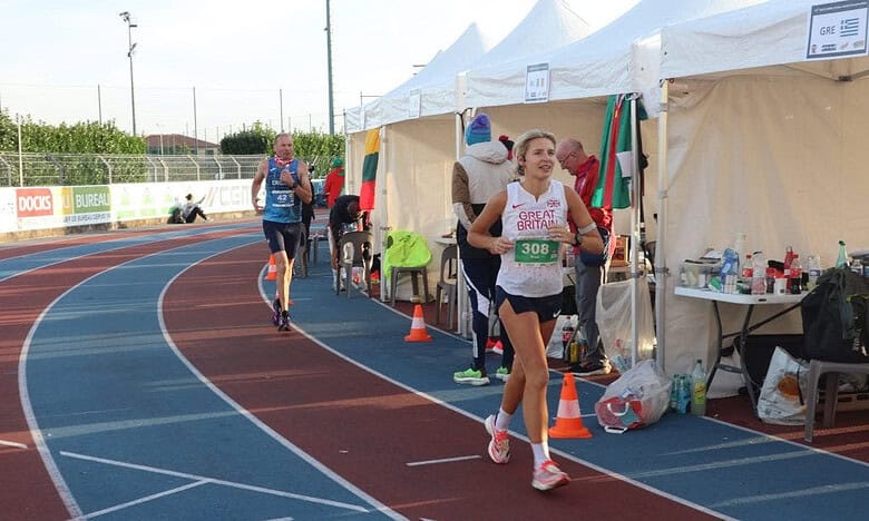Competitive female runner smiling at a track event in Great Britain, close to the finish line, during a marathon or race with support tents in the background, highlighting endurance and athletic achievement.