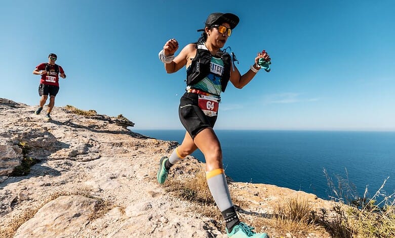 Elevated view of female trail runner navigating rocky coastal terrain during outdoor race, wearing hydration gear and sports attire, with her male running companion in the background, under clear blue sky.