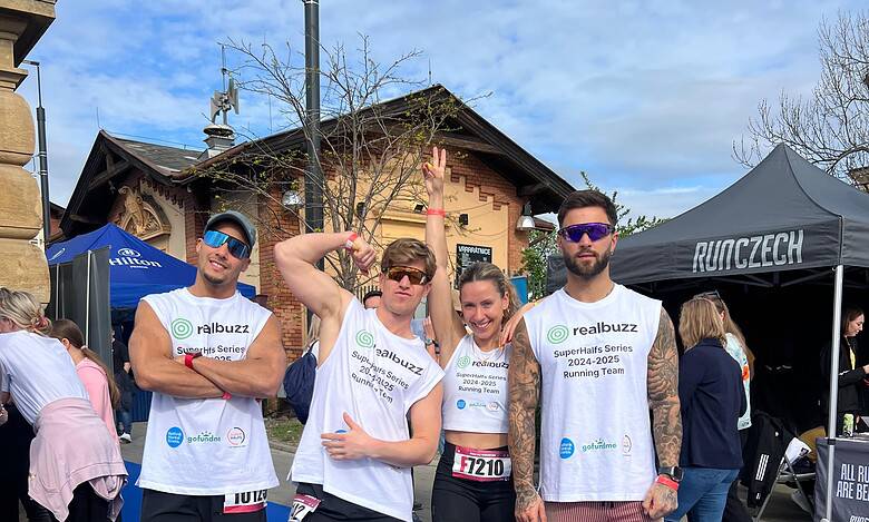 1. Group of runners at a marathon event wearing running team shirts, smiling and posing on a blue running mat after completing a race, with race bibs visible.
