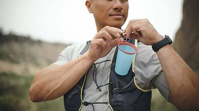 An athlete preparing for a run, holding a water bottle with a hydration tube, outdoors in a natural setting.