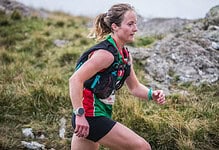An experienced female trail runner wearing a hydration pack, running through a rugged outdoor landscape with grass and rocks, showcasing endurance and outdoor adventure.