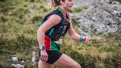 An experienced female trail runner wearing a hydration pack, running through a rugged outdoor landscape with grass and rocks, showcasing endurance and outdoor adventure.