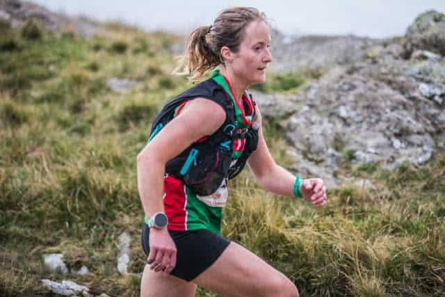 An experienced female trail runner wearing a hydration pack, running through a rugged outdoor landscape with grass and rocks, showcasing endurance and outdoor adventure.