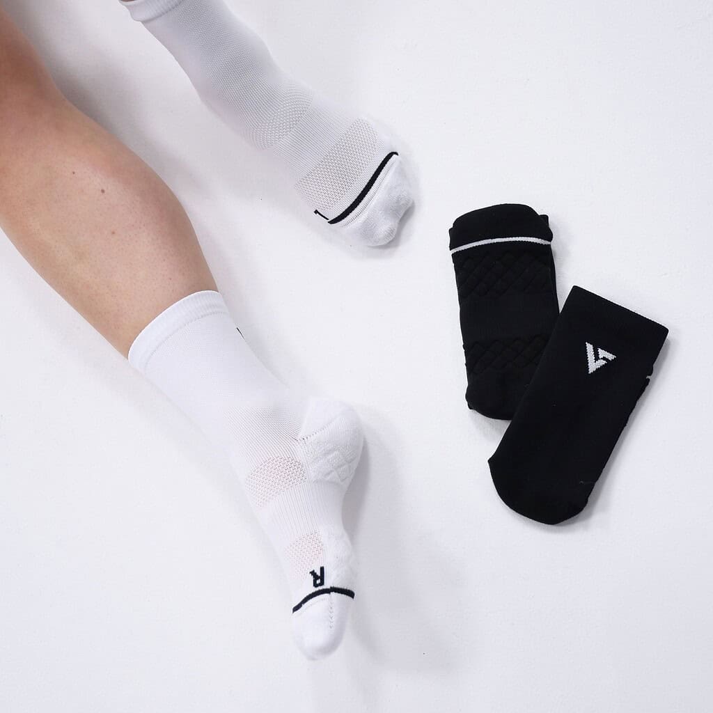 Close-up of a runner's legs wearing white Velocity Performance Crew Socks with two pairs of black socks nearby on a white background.