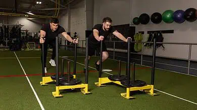 Two men performing sled push exercises in a gym, focusing on strength and endurance training for runners.