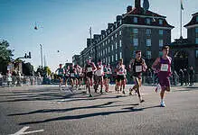 Running race with multiple athletes competing on a city street during daylight, with spectators and historic buildings in the background.