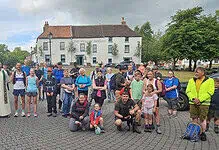 Running group gathering outdoors in a town square, diverse participants with backpacks and sportswear, some with dogs, preparing for a run or walk, under a partly cloudy sky.