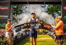 Triathlon athlete crossing the finish line at a race event, with supporters and event banners in the background, celebrating achievement and endurance.