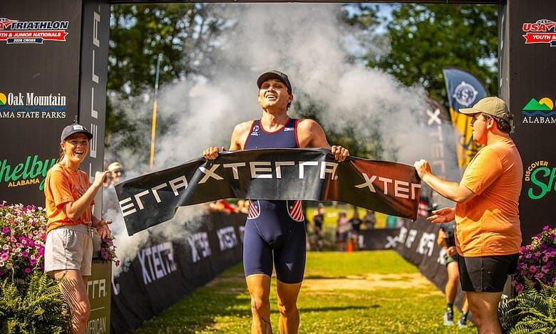 Triathlon athlete crossing the finish line at a race event, with supporters and event banners in the background, celebrating achievement and endurance.