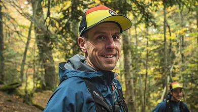 Male trail runner in colourful cap and waterproof jacket smiling in a forest during autumn.