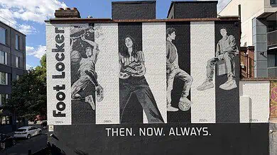 Black and white mural of four people sitting and standing, with 'Foot Locker' text and 'Then. Now. Always.' slogan on a city wall.