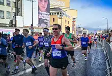 Group of marathon runners participating in a race on a city street, with spectators and urban buildings in the background.
