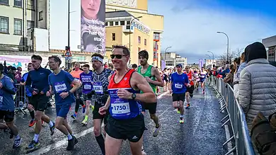 Group of marathon runners participating in a race on a city street, with spectators and urban buildings in the background.