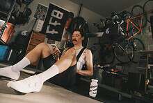 Man with a mustache and white socks sitting on the floor in a garage gym, looking at his smartphone, surrounded by cycling gear and equipment.