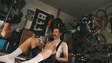 Man with a mustache and white socks sitting on the floor in a garage gym, looking at his smartphone, surrounded by cycling gear and equipment.