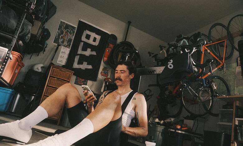 Man with a mustache and white socks sitting on the floor in a garage gym, looking at his smartphone, surrounded by cycling gear and equipment.