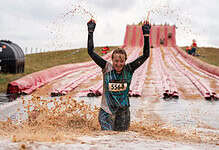 Female participant celebrating at Wolf Run obstacle course during muddy water challenge, wearing race bib 5564, with a large pink slide in the background.