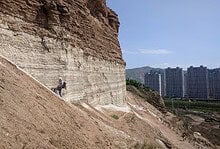 A person hiking along a steep, layered cliff face with a cityscape and mountains in the background.