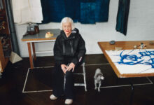 Elderly woman with white hair sitting in art studio with a small dog nearby, surrounded by fabric and artwork.