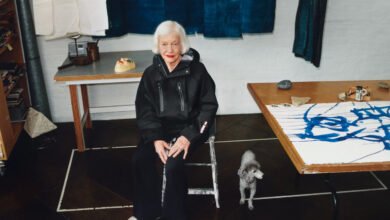 Elderly woman with white hair sitting in art studio with a small dog nearby, surrounded by fabric and artwork.