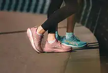 Two runners wearing pink and turquoise running shoes standing on a city sidewalk near a railing, ready for a run.
