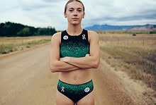 Young female runner standing confidently on a rural dirt trail with open fields and mountains in the background, wearing running gear, under cloudy skies.
