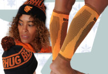 Person wearing orange knee-high socks and two knit beanies with pom-poms, set against a pale blue background with snowflake patterns.