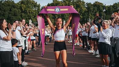 Female runner celebrating victory at a marathon finish line with a large purple ribbon, surrounded by cheering supporters on a sunny day.