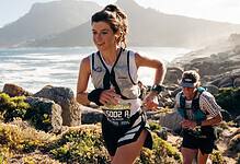 Two female trail runners competing on a rugged coastal trail with mountains and ocean in the background.