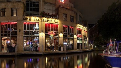Night view of Hard Rock Cafe Amsterdam illuminated with neon lights, reflecting on the canal water, with boats docked nearby.