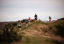 Group of runners participating in trail running on a scenic outdoor path with grassy hills and bushes under a cloudy sky.