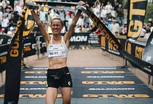 Female runner crossing the finish line with arms raised in victory at a sporting event, wearing a white sports top and black shorts, crowd in background.