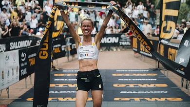 Female runner crossing the finish line with arms raised in victory at a sporting event, wearing a white sports top and black shorts, crowd in background.