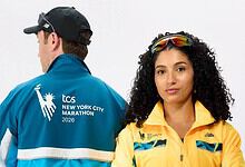 Two runners pose for a studio shot: a man in a blue jacket with TCS New York City Marathon 2026 logo on the back, and a woman in a yellow jacket with reflective sunglasses on her head, against a white.