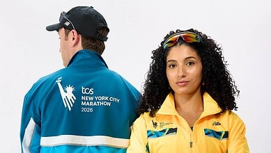 Two runners pose for a studio shot: a man in a blue jacket with TCS New York City Marathon 2026 logo on the back, and a woman in a yellow jacket with reflective sunglasses on her head, against a white.